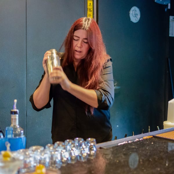 A bartender with long red hair shakes a cocktail shaker behind a bar with bottles and glasses.