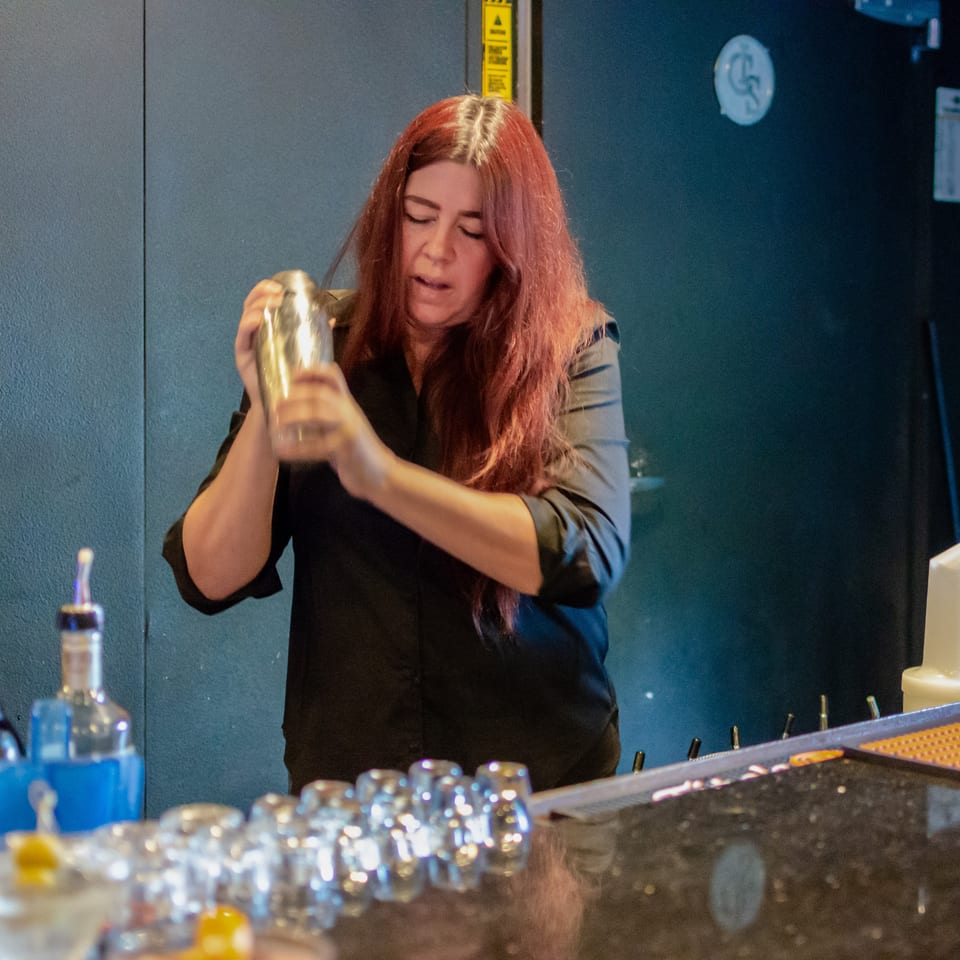 A bartender with long red hair shakes a cocktail shaker behind a bar with bottles and glasses.