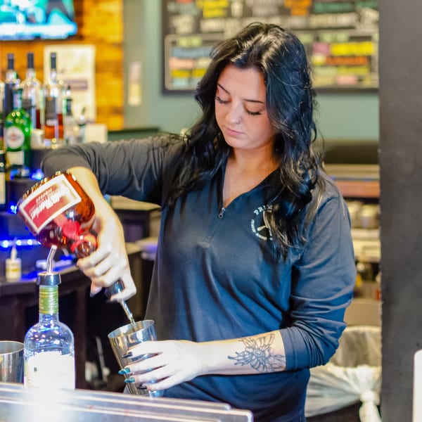 Bartender in a black shirt pouring liquor from a bottle into a cocktail shaker at a bar.