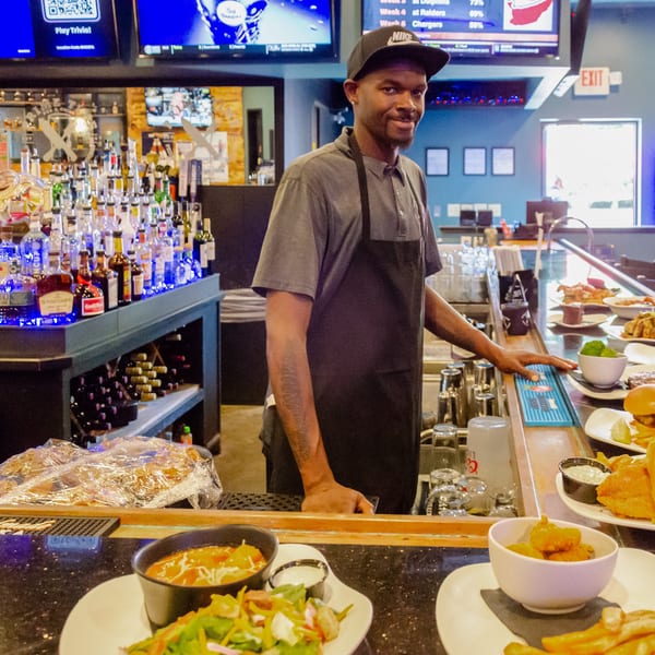 A bartender in a black apron smiles behind a bar with many liquor bottles and plates of food.