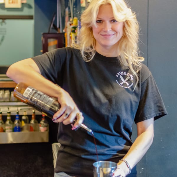 Blonde bartender in a "Prime Time" shirt pouring a drink at the bar.