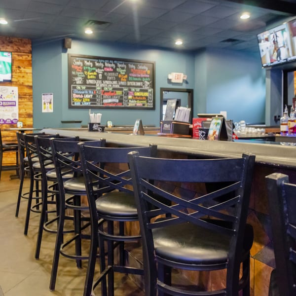 Empty bar area with black stools, a chalkboard menu, and TVs showing sports.