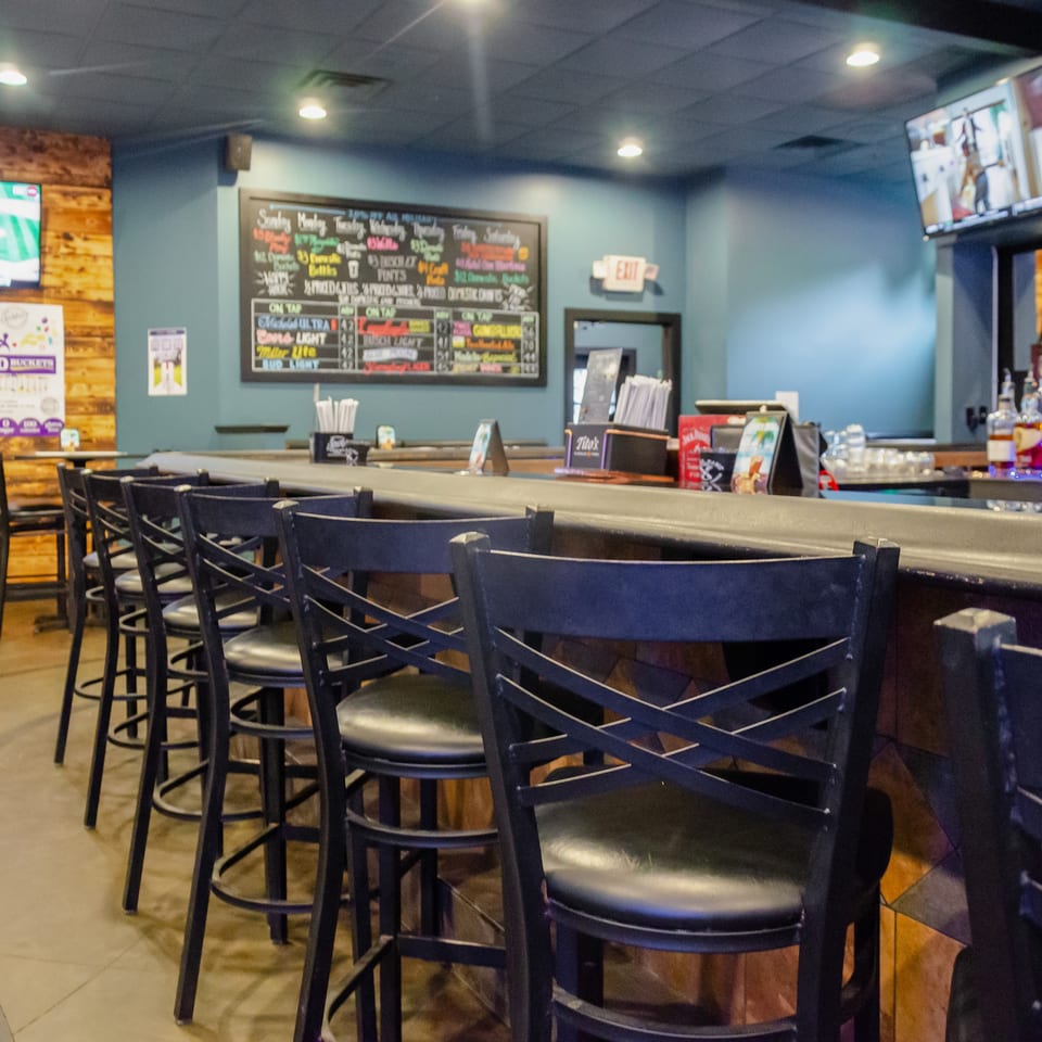 Empty bar area with black stools, a chalkboard menu, and TVs showing sports.