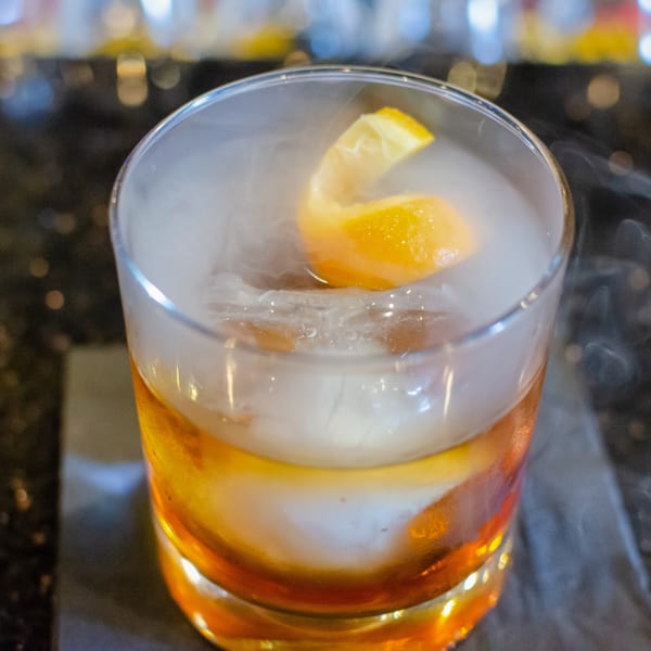 Smoking amber cocktail with an orange peel and ice on a dark bar surface.