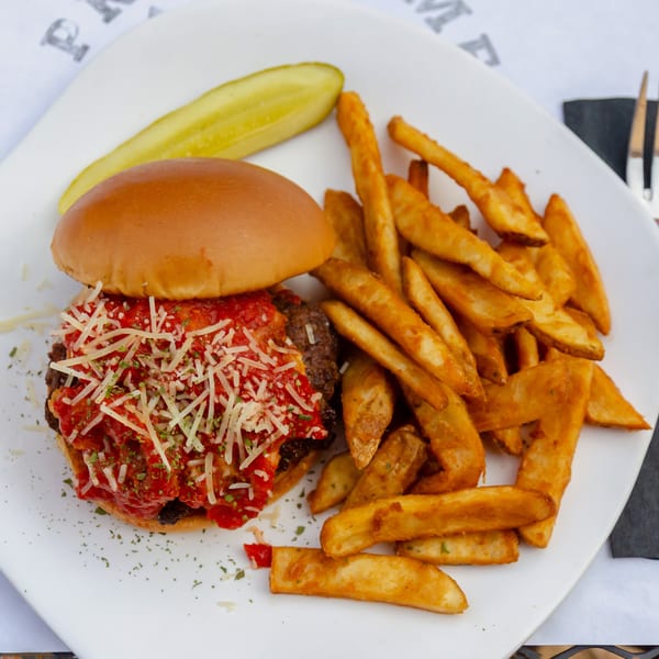A burger topped with marinara sauce and shredded cheese, served with fries and a pickle.