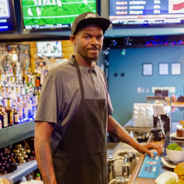 Man in a Nike cap and apron stands behind a bar with liquor bottles, food, and TVs.