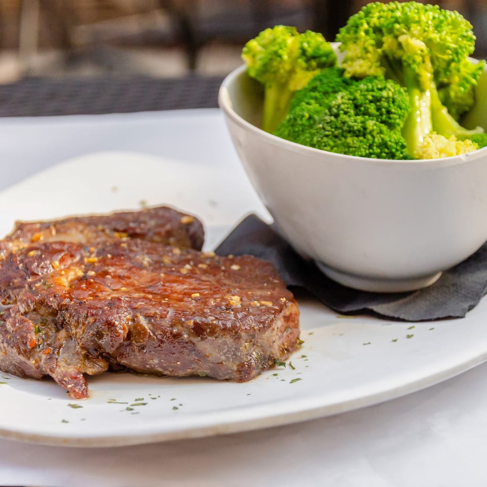 Grilled steak with herbs and a side of steamed broccoli in a white bowl on a white plate.